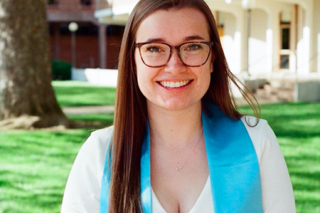Lauren smiling in front of a building wearing glasses and a graduation sash